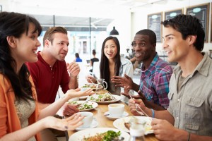 Diverse group of individuals sits around a table talking and eating. 