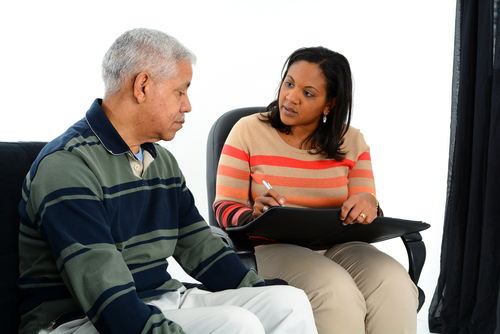 Two people, sitting and having a conversation. One person is an older man with short, gray hair. The other person is a woman with dark hair; she is holding a pen and notebook.