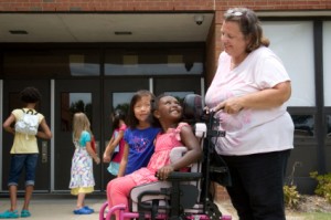 A group of people outside a school. There are three school-aged children in the background. The focus of the picture is an adult woman pushing a girl in a wheelchair; they are looking at each other and smiling.