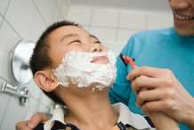 A young person with short black hair and shaving cream on their face is getting help to shave.