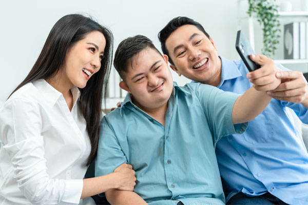 Three people sitting closely together who are smiling while taking a selfie. The person on the left is a woman, in the middle is a young man with a disability, on the right is another man.