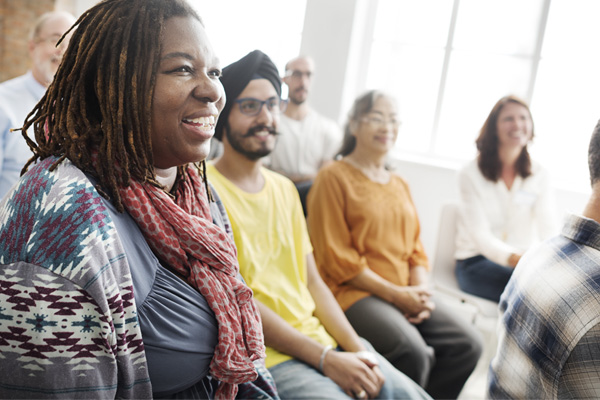 A group of ethnically diverse people sitting together in a casual, friendly setting, engaged in conversation. The focus is on a woman with dark skin and dreadlocks, who is smiling and wearing a patterned shawl.