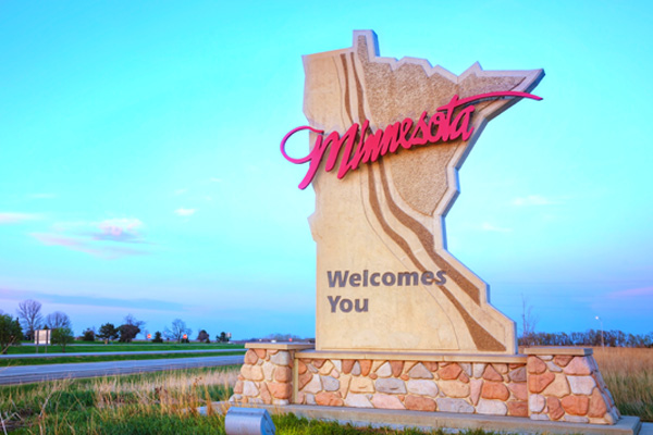 A sign for the state of Minnesota. The sign is shaped like the state itself and has the word "Minnesota" written in large, red cursive letters across the top. Below that , it says "welcomes you" in smaller, black print. The sign is mounted on a stone base.