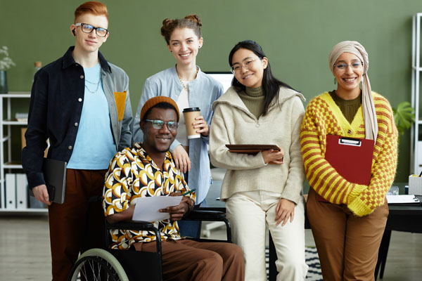 A group of five young, ethnically diverse, people in an office smile at the camera. Four are standing and one person is in a wheelchair.