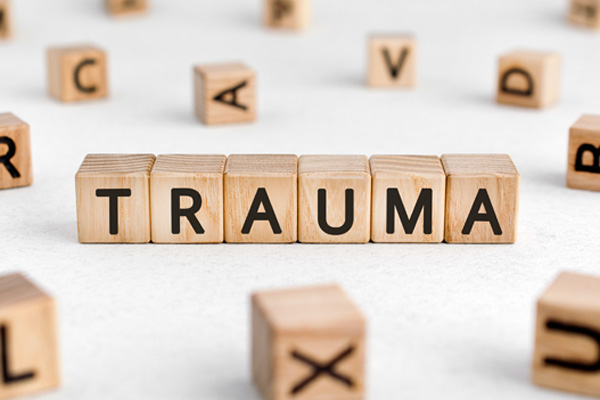 Wooden blocks with letters on them, arranged to spell the word "trauma" vertically. The blocks are placed on a light-colored surface, and there are other scattered blocks with letters in the background.