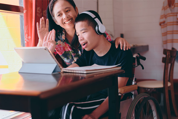 A young boy in a wheelchair is wearing headphones. He is sitting at a table with an open notebook and a tablet in front of him. A woman, who is a caregiver, is sitting behind him smiling and waving at the tablet.