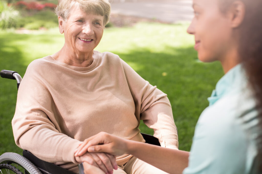 An older white woman sits outside in a wheelchair holding hands with a younger woman who faces her. They are looking at one another and smiling. It is sunny outside.