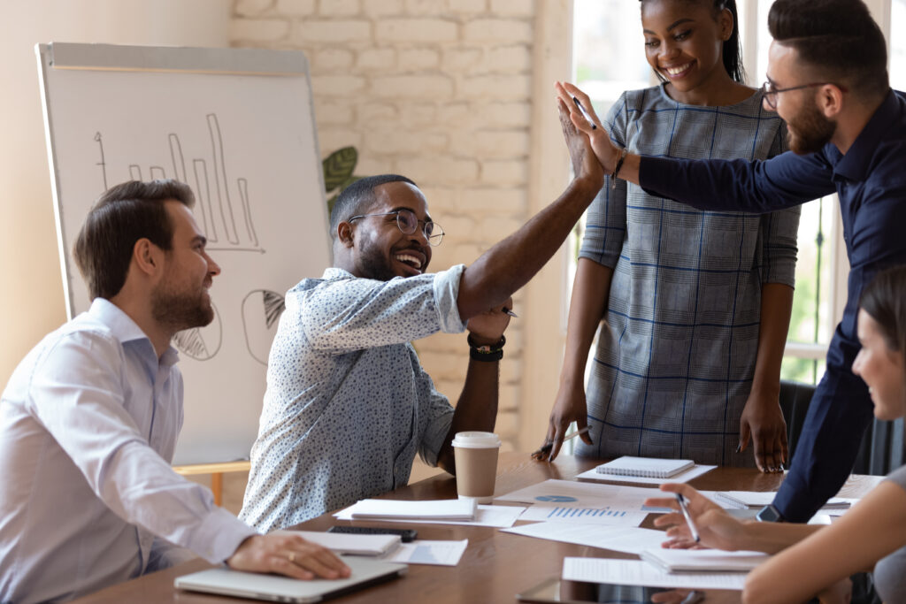 An ethnically diverse group of young professionals sits at a meeting space in an office. They appear happy. Two young men high five while the others watch.