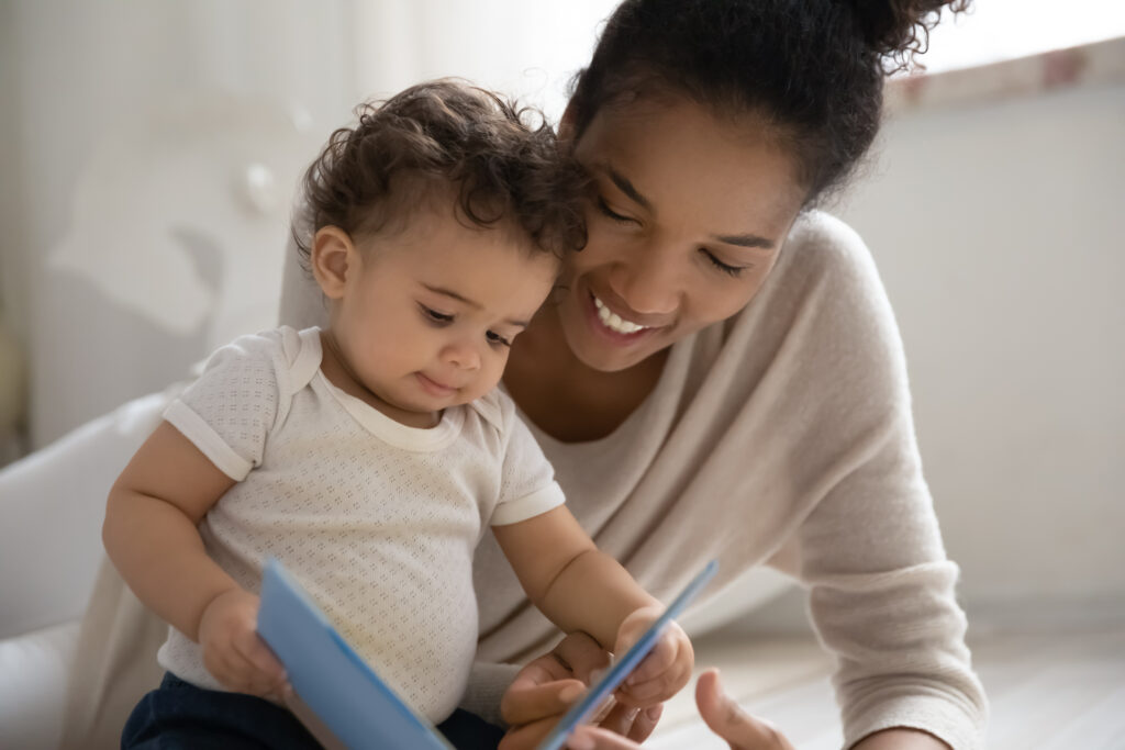 A young black mother smiles while she sits with her baby, who holds open a blue book. They are both looking at the book.