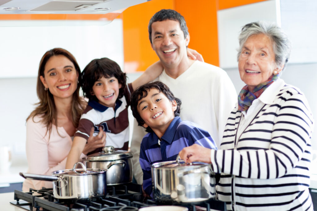 A multi-generational family in a kitchen. The family includes a woman with long brown hair, two young boys with dark brown hair, a man with short brown hair, and an older woman with short gray hair. They are all smiling.