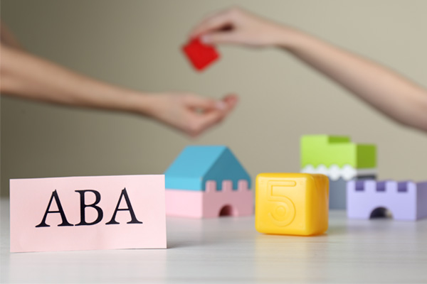 Colorful toy blocks are arranged in a row. The blocks are various shapes and colors. Next to the blocks there is a piece of paper with the letters "ABA" written on it in large, black font. Two arms are visible, one holding a red block and the other reaching out.