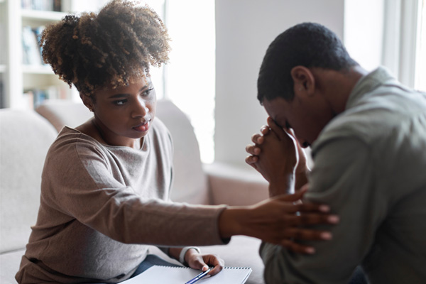 Two people interacting closely in a living room. One person is a woman with curly hair; she is leaning towards the other person, who is a man. He is holding his hands up to his face. The woman is reaching out to touch the man's arm.