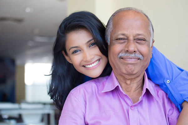 An older man and a younger woman are smiling. The man has short gray hair, a mustache, and is wearing a purple shirt. The woman, who is standing behind him with her arms around his shoulders, has long dark hair.