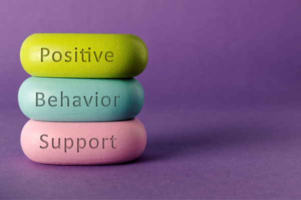 Three colorful, pill-shaped objects arranged vertically against a purple background. Each object has a word written on it. The top object is green and has the word "positive." The middle object is blue and has the word "behavior." The bottom object is pink and has the word "support." These words together form the phrase "positive behavior support."