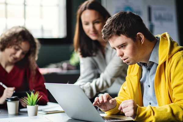 Three people sitting at a table, engaged in collaborative work. The person in the foreground is a young man with down syndrome. He is looking intently at a laptop in front of him. Behind him, there are two women who are also focused on the work at hand.