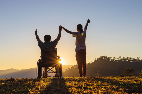 Two people outdoors during a sunset. One person is standing and holding the hands of another person who is sitting in a wheelchair. Both have their arms raised in a joyful manner. The background is a scenic view.