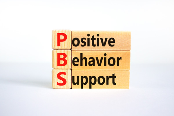 Wooden blocks arranged to spell out the words "Positive Behavior Support." The blocks are stacked vertically, with each word on a separate block. The first letters of each word are in red, while the rest of the letters are in black. The words are aligned so that the first letters form the acronym "PBS."