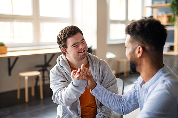 Two men interacting in a friendly manner. The first man, who has short dark hair and down syndrome, is leaning forward and smiling, He is reaching out to fist bump the other man. The other man, who has a dark complexion, beard, and short curly hair, is looking at the first man.