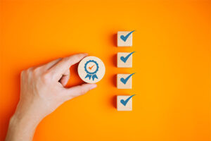 A hand holding a circular object with a symbol of a blue ribbon with an orange check mark in the middle. Next to the hand there are four square tiles arranged in a vertical row. Each tile has a blue check mark in it. The background is bright orange.