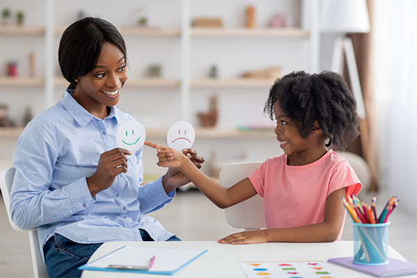 A woman and a young girl engaging in playful activity. The woman, who has dark skin, is holding a paper craft with a smiley face. The child, who also has dark skin, is reaching up to touch the craft. They are both smiling.