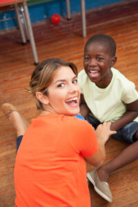 A teacher in an orange shirt looks over her shoulder at the camera smiling. She is holding the hands of a young black student who is looking at her smiling. They are both sitting on the floor in a classroom.