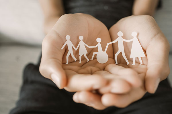 A pair of hands gently cupping a group of paper cut-out figures. The figures are holding hands and appear to represent different types of people, including one in a wheelchair.