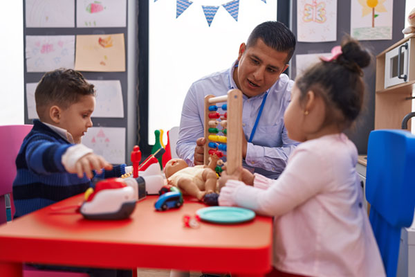 A man, who is a teacher, and two young children sit at a table, engaged in play. The teacher is smiling and interacting with the children.