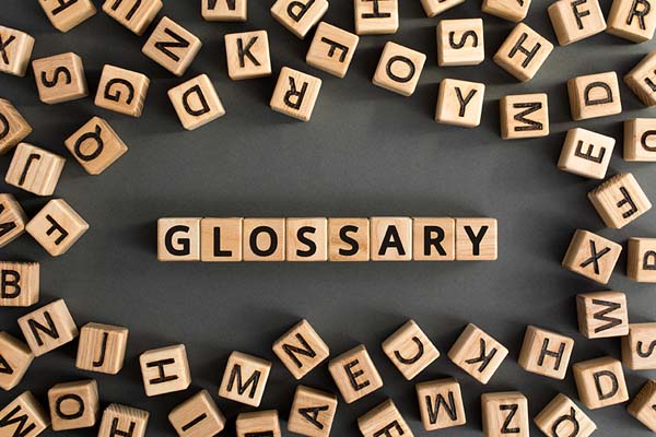 A collection of wooden letter blocks arranged on a dark surface. The blocks spell out the word "glossary" vertically in the center. Surrounding the word are numerous other blocks scattered randomly, each displaying a single letter.