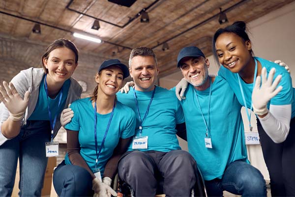 A group of ethnically diverse people posing together, waving at the camera, and smiling. They're all wearing matching blue shirts and white gloves. They're in a casual setting.