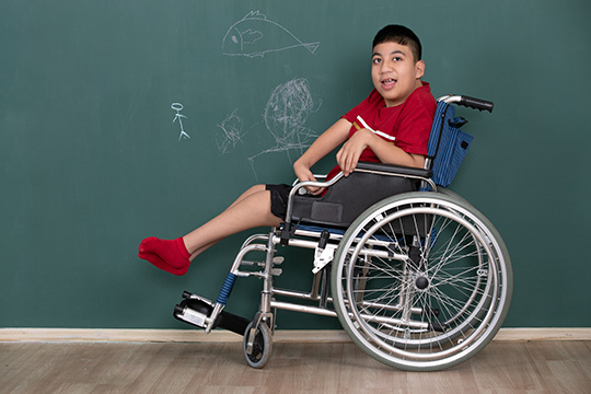 A boy in a wheelchair sits in front of a chalkboard with doodles drawn on it.