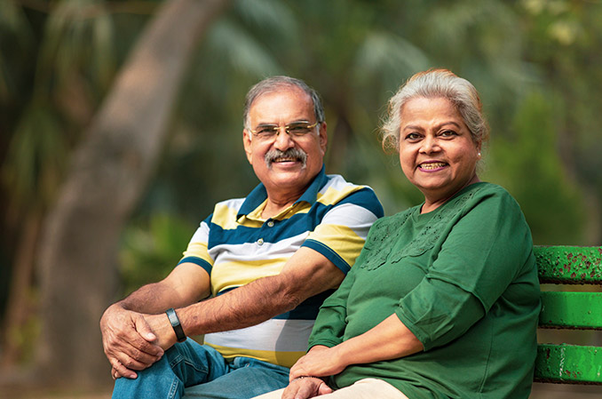 An older couple is smiling, while sitting on a green bench outside.