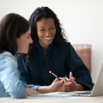 Two women sitting side by side smiling and looking at laptop