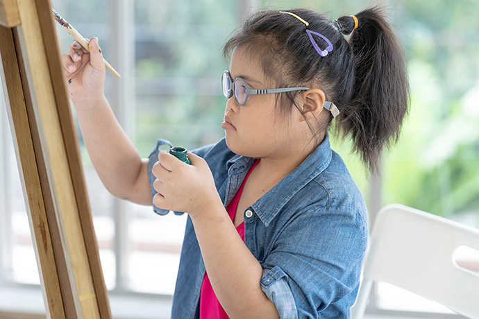 A young girl with down syndrome paints at an easle.