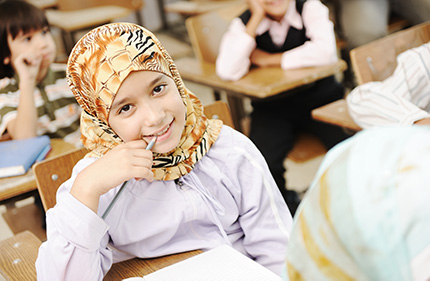 Children in a classroom, the focus is on a girl wearing a hijab as she smiles and looks at the camera.