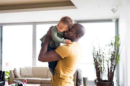 A black father holds his daughter up near his face, he is smiling, she is looking away from him smiling.