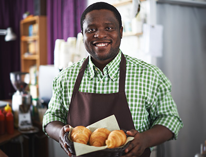 A man wearing a green and white checkered shirt and brown apron and has a dark complexion. He is holding some croissants and smiling.