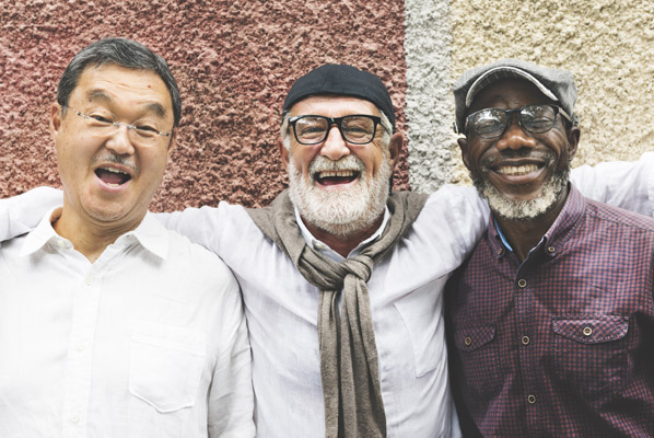 Three older men are smiling and appear happy. One man has short, dark hair, another man has a white beard, and the last man has a dark complexion and a white beard. They're all wearing glasses.