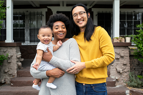 A young, ethnically diverse man and woman stand in front of a house holding their baby. All of them are smiling and look happy.