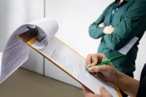 A person writing on a notepad with a green pen. In the background there is another person wearing a green jacket, but they are out of focus.