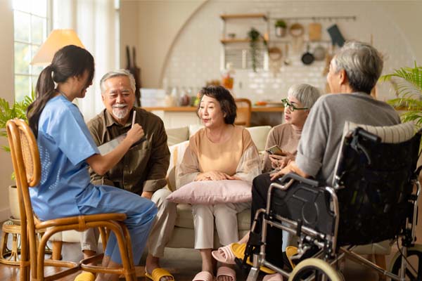 A group of people in a cozy living room. There are five people including a nurse in a blue uniform, who is engaging with the others, an older man, two older women, and another older person in a wheelchair.