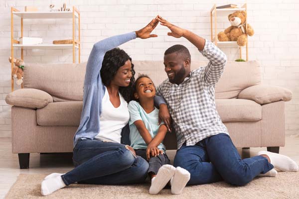 A happy family of three sitting on the floor in a living room. The family consists of a mother, father, and young child. The mother and father are sitting on either side of the child, and they have their arms raised above the child's head, forming a hear shape with their hands.