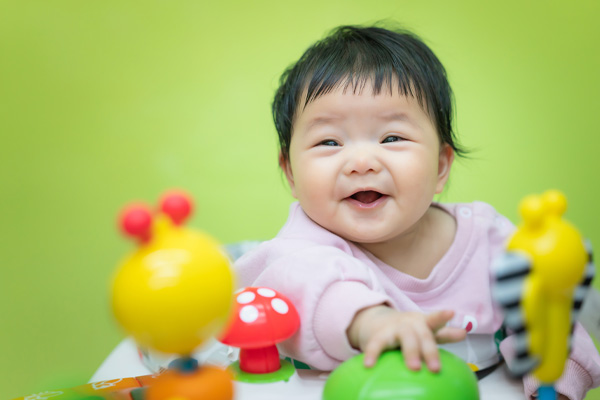 An image of a baby with short dark hair, wearing a light pink outfit. The baby is smiling and appears to be playing with some colorful toys.