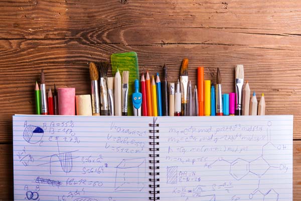 An image of an open spiral notebook on a wooden surface. The notebook is filled with handwritten notes and drawings, including geometric shapes and some text. To the right of the notebook, there is a collection of various colored pencils, pens, and markers arranged neatly.