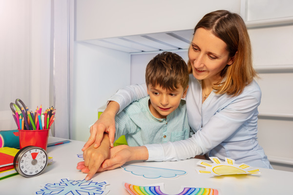 A woman and a young boy sitting at a table. The woman is providing hand over hand support while they point to colorful weather drawings on the table in front of them, including a snowflake, rainbow, cloud, and sun.