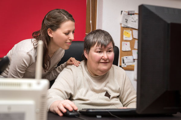 Two people in front of a computer. One person, a woman with short hair, is sitting and looking at the computer screen. The other person is a woman with long hair, standing behind, leaning in and looking at the screen as well.