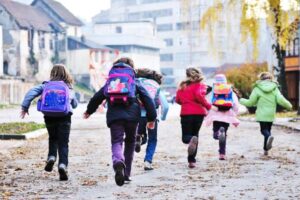 A group of children walking away from the camera on a dirt path. They are wearing colorful backpacks and are dressed in winter clothing, such as jackets and boots. The background includes some buildings and trees.