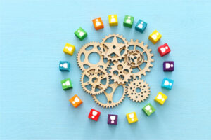 A collection of wooden gears arranged in a circular pattern in the center. Surrounding the gears is another circle made up of small, colorful blocks. Each block has a different color and features a white icon of a person on it.