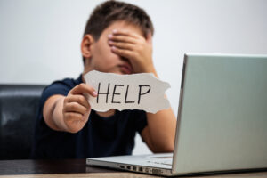 A boy is covering his eyes with his hand while sitting at a table with a laptop open. He holds a sign that says help.