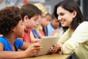 A woman is interacting and smiling at a young girl holding a tablet. In the background, there are other children, slightly out of focus, including a boy who is engaged in similar activities.